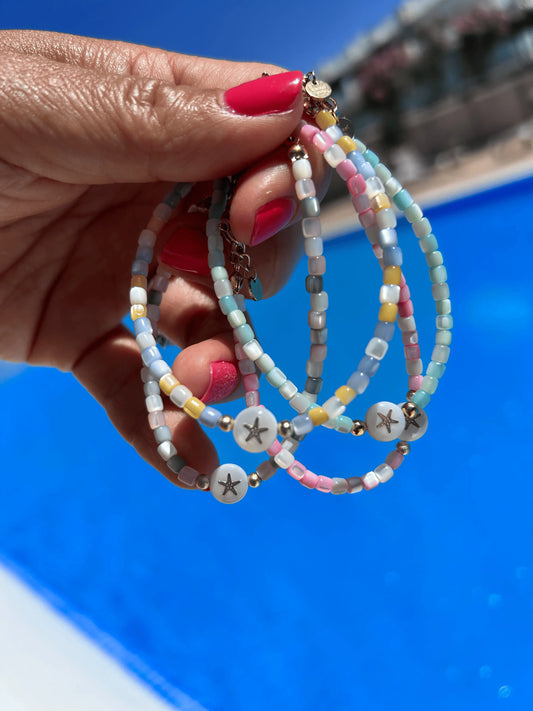 Bracelets en perles nacre colorées bleu, rose, jaune, blanc et gris, ornés d'une perle à motif étoile de mer en nacre blanche en son centre. Bijoux sur fond d'une piscine extérieure.