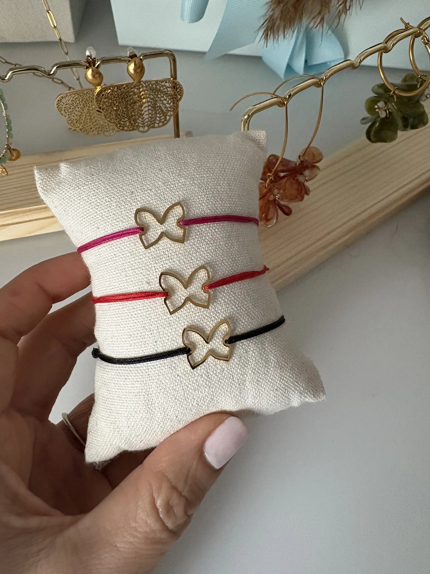 Trois bracelets cordon en nylon tressé de couleurs noir, rouge et fuchsia, ornés d'un papillon en acier inoxydable doré. Bijoux présentés sur un mini-coussin en coton beige. En arrière plan, des boucles d'oreilles papillon en acier odré et des boucles d'oreilles hameçon et créoles dorées ornées de pétales en acrylique colorées.