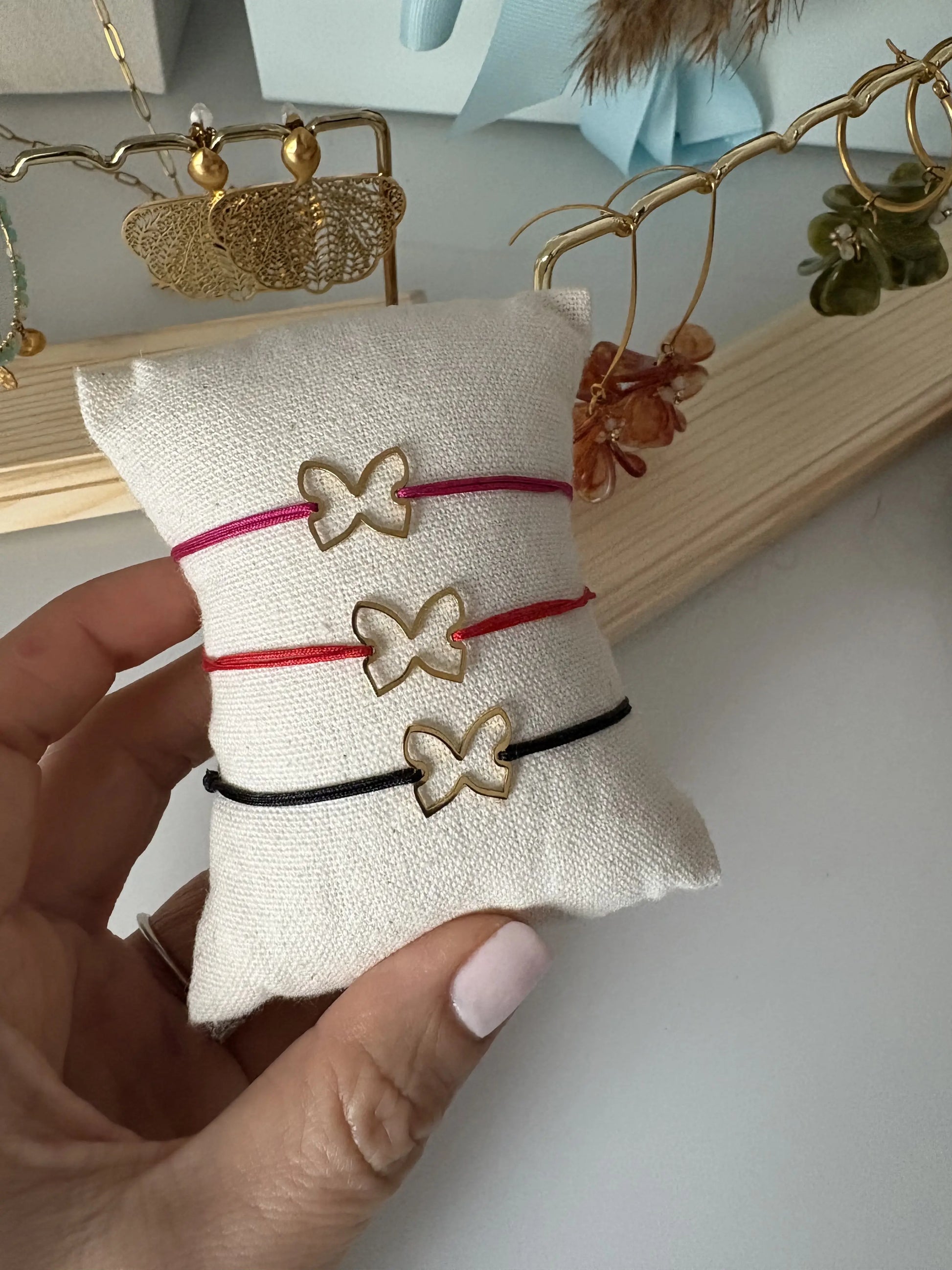 Trois bracelets cordon en nylon tressé de couleurs noir, rouge et fuchsia, ornés d'un papillon en acier inoxydable doré. Bijoux présentés sur un mini-coussin en coton beige. En arrière plan, des boucles d'oreilles papillon en acier odré et des boucles d'oreilles hameçon et créoles dorées ornées de pétales en acrylique colorées.