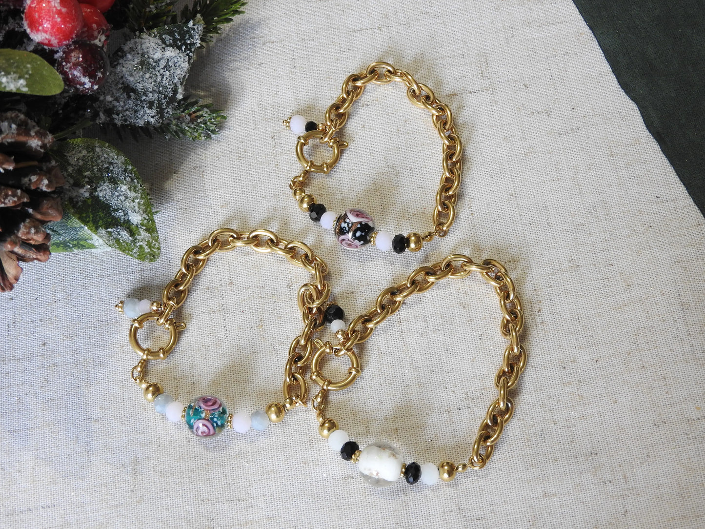 Trois bracelets posés à plat sur une nappe beige décoré de feuilles de sapin. Bijoux mi-chaîne grosses mailles et mi-perles boules en verre au motif floral de couleurs noir, verte et blanche.