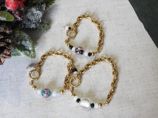 Trois bracelets posés à plat sur une nappe beige décoré de feuilles de sapin. Bijoux mi-chaîne grosses mailles et mi-perles boules en verre au motif floral de couleurs noir, verte et blanche.
