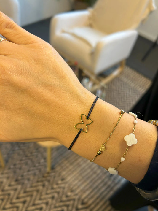 Bracelets portés au poignet. Un bracelet cordon en nylon tressé noir orné d'un papillon en acier inoxydable doré. Un bracelet chaîne fine avec une perle coeur plate à effet diamanté et un bracelet chaîne en acier doré et perles de verre blanche et perles trèfle en nacre blanche.