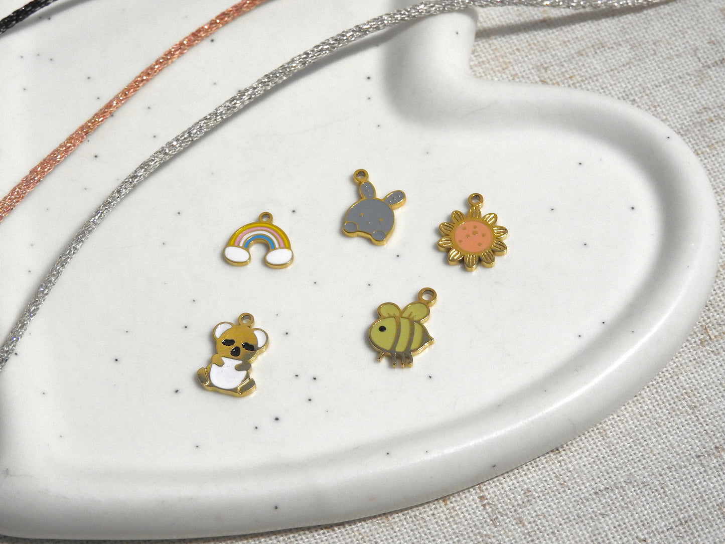 Pendentifs breloques disposés sur une coupelle en céramique blanche. Bijoux pour enfants lapin, tournesol, arc-en-ciel, koala et abeille.