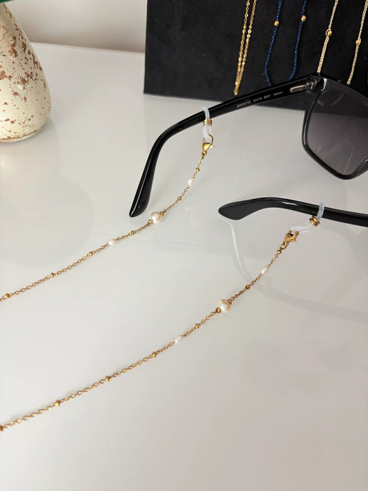 Bijou chaîne de lunettes en acier inoxydable doré et perlée, ornée de perles d'eau douce blanche. Bijou monté sur les branches de lunettes de soleil noires.