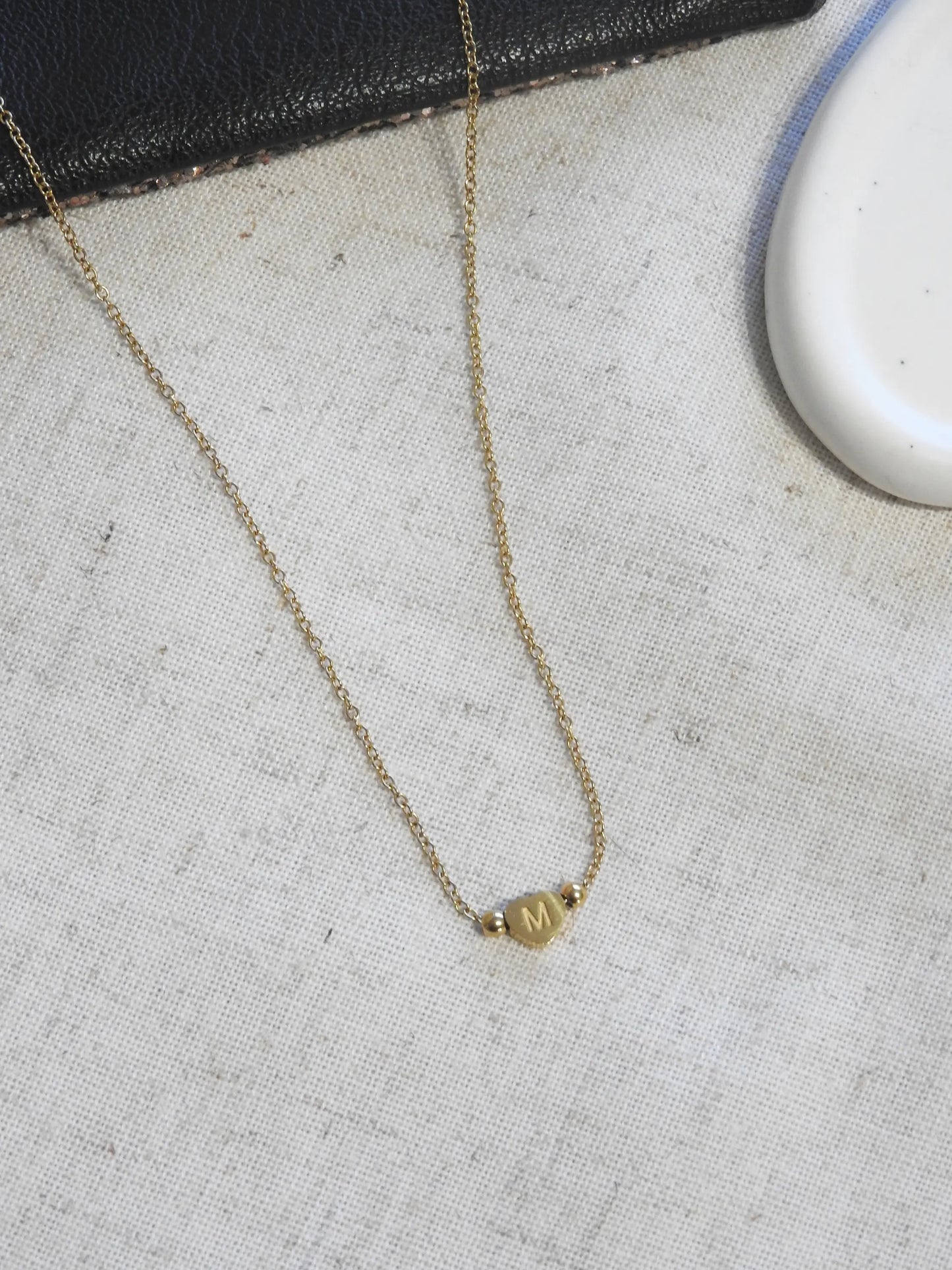 Collier chaîne en acier inoxydable doré posé à plat sur une nappe beige et pochette noire. Bijou orné d'une perle plate en forme de coeur, gravé de la lettre M.