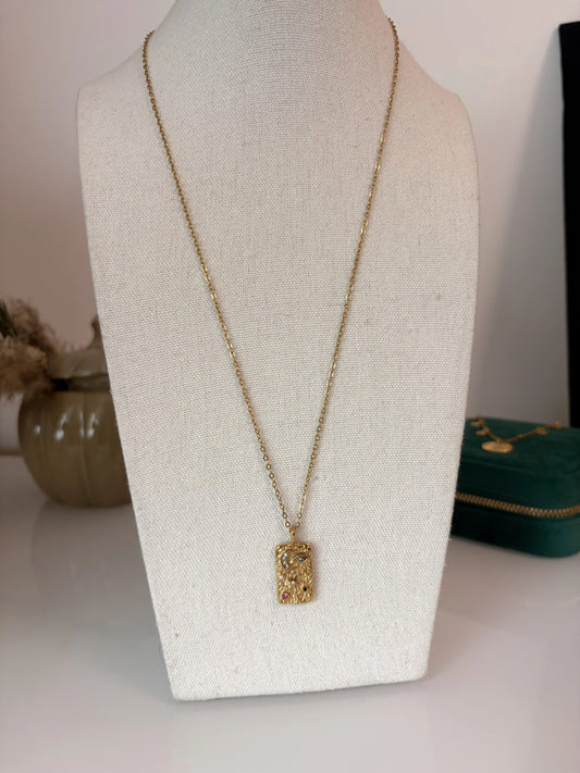 Long collier chaîne en acier inoxydable doré et orné d'un pendentif rectangle avec des symboles lune, soleil et oeil protecteur strass. Bijou présenté sur un buste en coton beige.