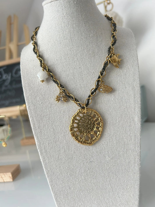 collier chaine en acier inoxydable doré et orné d'un cordon tissé noir et doré. Bijou orné de 5 charms disposés sur la chaine avec un coquillage en céramique blanc, un crabe, un coquillage fossilisé au centre, un coquillage allongé et un poisson. Bijou sur un présentoir buste en coton beige.