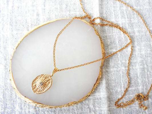 Collier sautoir chaîne en acier inoxydable doré avec un pendentif médaille de forme ovale orné d'un soleil en relief avec une pierre en oxyde de zirconium.  Bijou posé sur une plaque ronde en acrylique blanche à bordure dorée.
