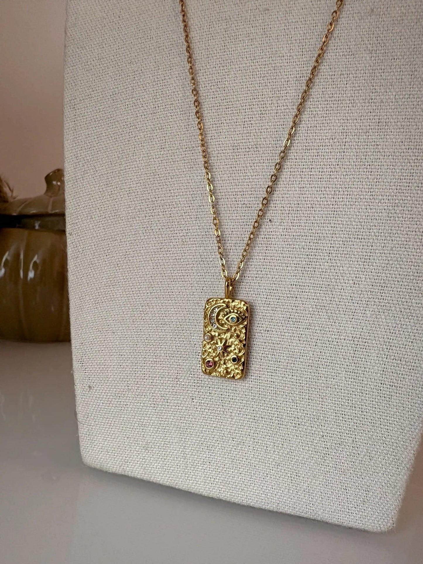 collier chaîne en acier inoxydable doré à pendentif rectangle orné de symboles forts lune, soleil et oeil protecteur strass. Bijou présenté sur un buste en coton beige.