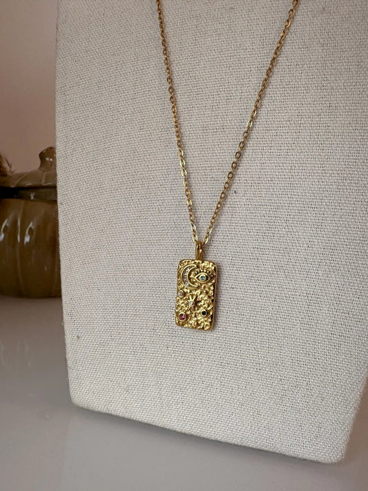 collier chaîne en acier inoxydable doré à pendentif rectangle orné de symboles forts lune, soleil et oeil protecteur strass. Bijou présenté sur un buste en coton beige.