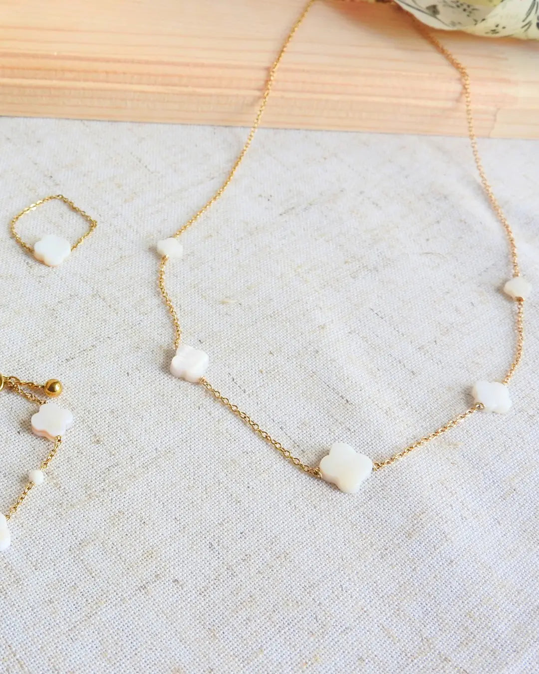 collier chaîne en acier inoxydable doré orné de perles en forme de trèfle en nacre blanche. Bague chaînette ornée d'une perle trèfle en nacre blanche. Bijoux posés sur une nappe beige en coton et plaque en bambou.