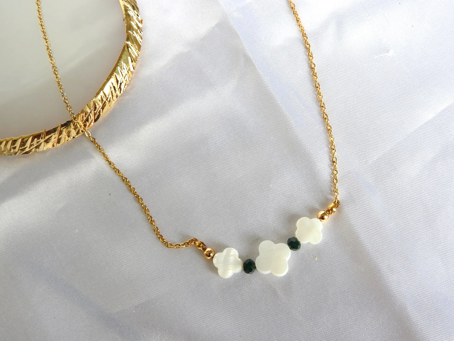 Collier chaîne en acier inoxydable doré orné de trois perles trèfle en nacre blanche et de perles de verre de couleur verte. Bijou posé sur une nappe blanche.