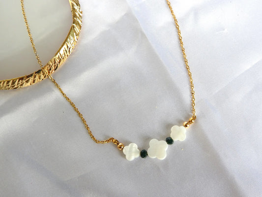 Collier chaîne en acier inoxydable doré orné de trois perles trèfle en nacre blanche et de perles de verre de couleur verte. Bijou posé sur une nappe blanche.