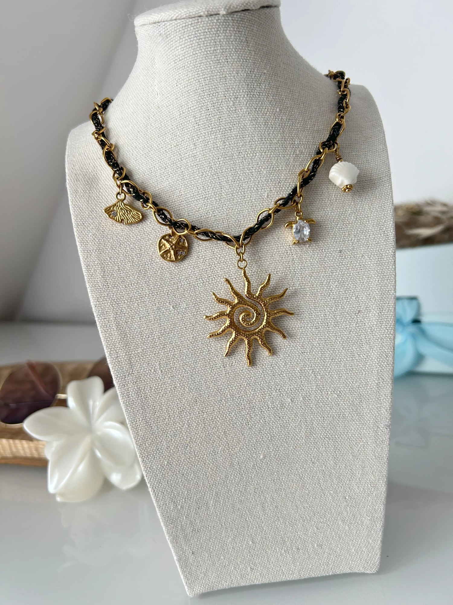 collier chaine en acier inoxydable doré et orné d'un cordon tissé noir et doré. Bijou orné de 5 charms disposés sur la chaine avec un coquillage allongé, une étoile de mer, un grand soleil au centre, une tortue de mer et un coquillage en céramique blanc. Bijou sur un présentoir buste en coton beige, posé sur une table décorée d'une pince fleur blanche et lunette solaire.