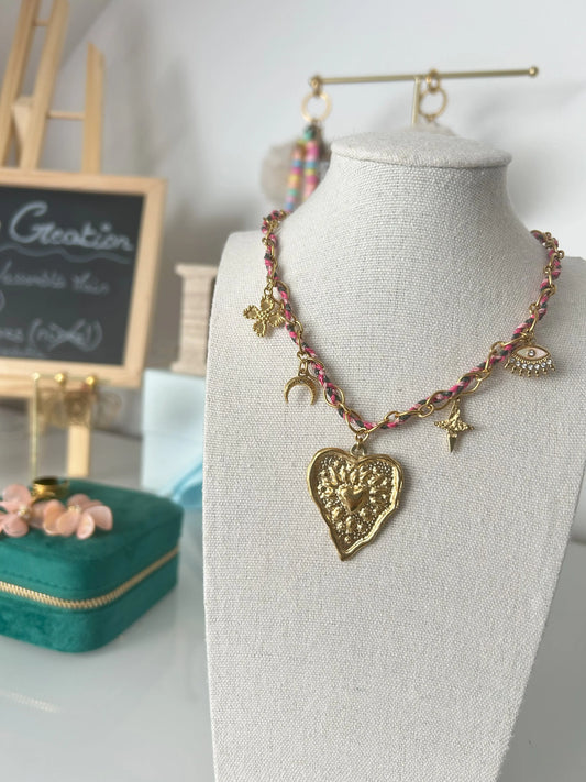 collier chaine orné d'un cordon de couleur rose, gris et marron. Bijou orné de 5 charms disposés sur la chaine avec un trèfle, une corne demi-lune, un grand coeur au centre, une étoile du nord martelé et un oeil protecteur.