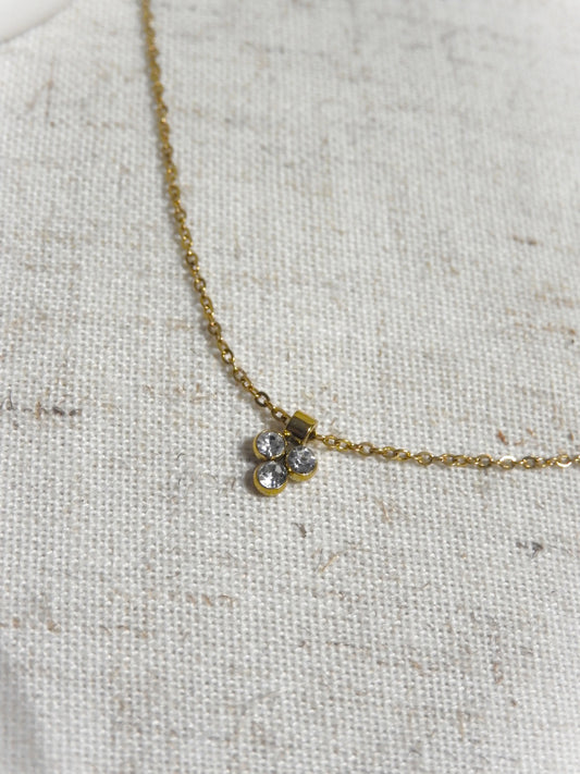 collier fine chaîne en acier inoxydable doré, orné d'un pendentif 3 pierres en oxyde de zirconium et acier inoxydable doré. Bijou posé à plat sur une nappe beige en coton.