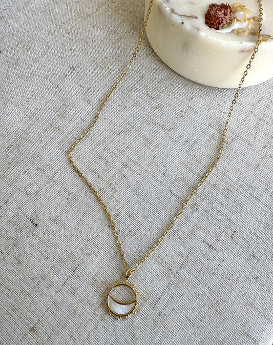 collier chaine en acier inoxydable doré orné d'un pendentif soleil évidé avec un formant un croissant de lune en nacre blanche. Bijou posé sur une nappe beige décorée d'une bougie fleurie