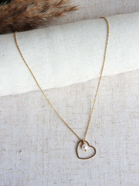 collier chaîne fine avec un pendentif coeur évidé et orné d'une perle d'eau douce. Bijou en acier inoxydable doré posé sur une nappe beige décorée d'une brindille.