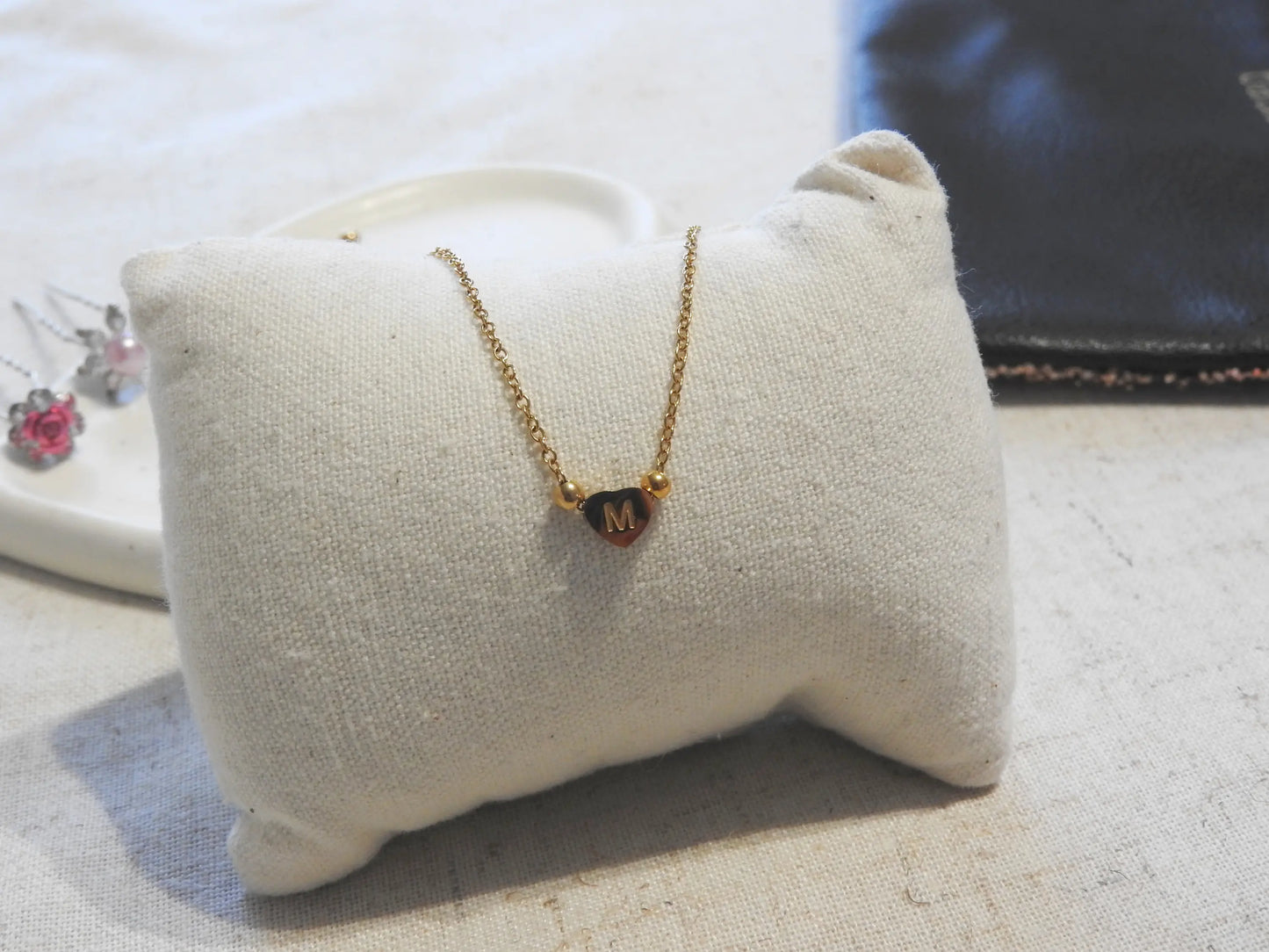 collier à chaîne fine orné d'une perle plate en forme de coeur et gravée de la lettre M. Bijou en acier inoxydable doré posé sur un mini-coussin beige.