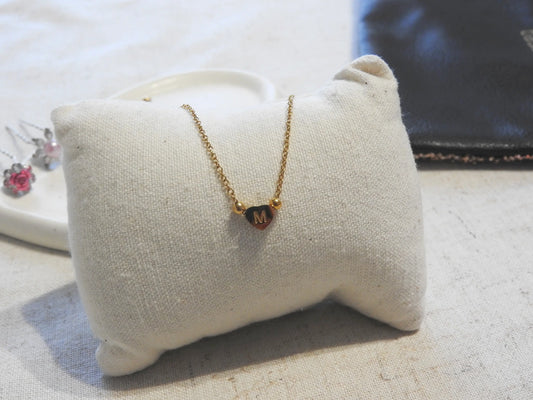 collier à chaîne fine orné d'une perle plate en forme de coeur et gravée de la lettre M. Bijou en acier inoxydable doré posé sur un mini-coussin beige.