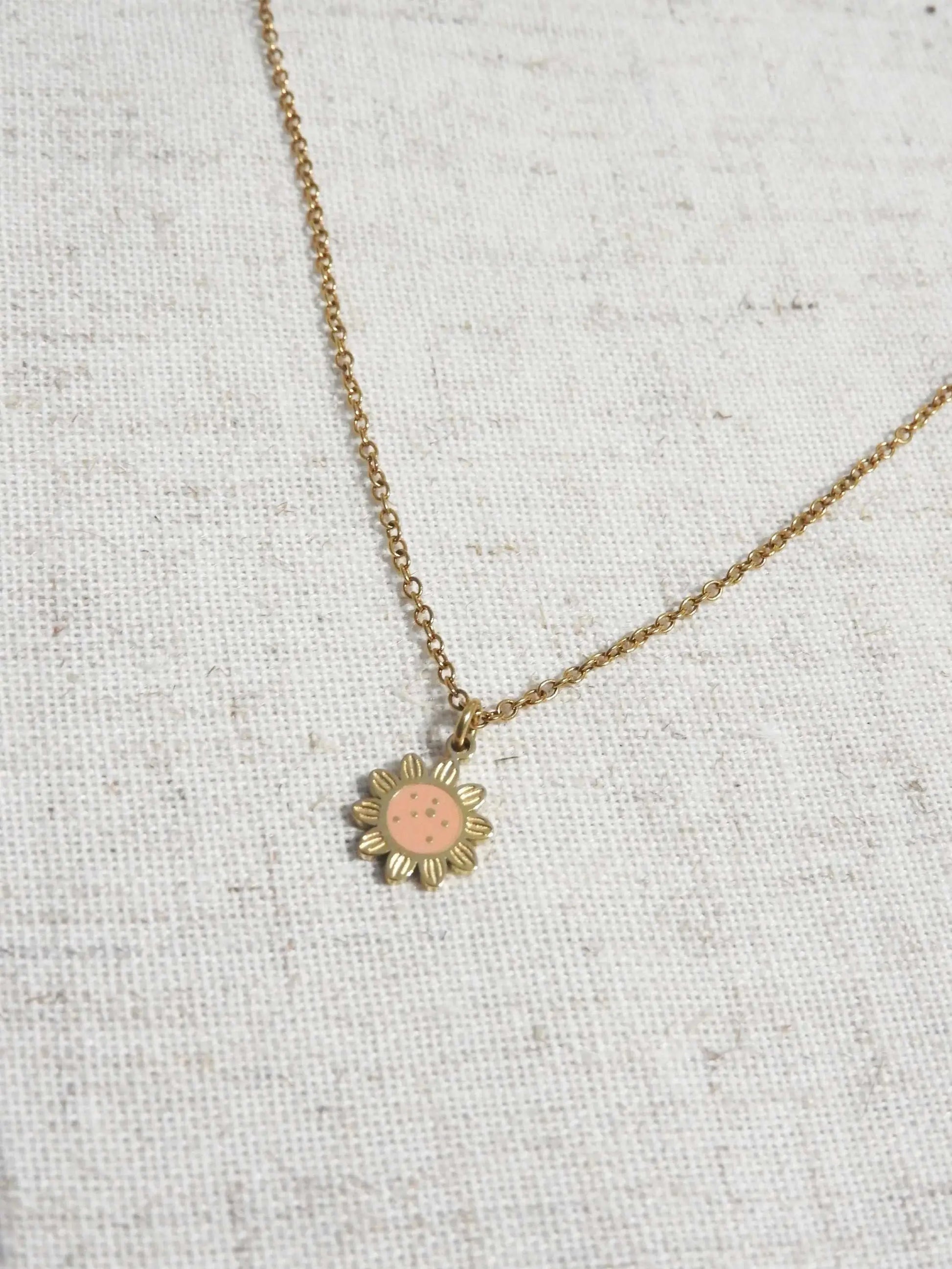 Collier pour enfant à chaîne fine orné d'un pendentif tournesol saumon en résine époxy et acier inoxydable doré. Bijou posé à plat sur une nappe beige en coton.