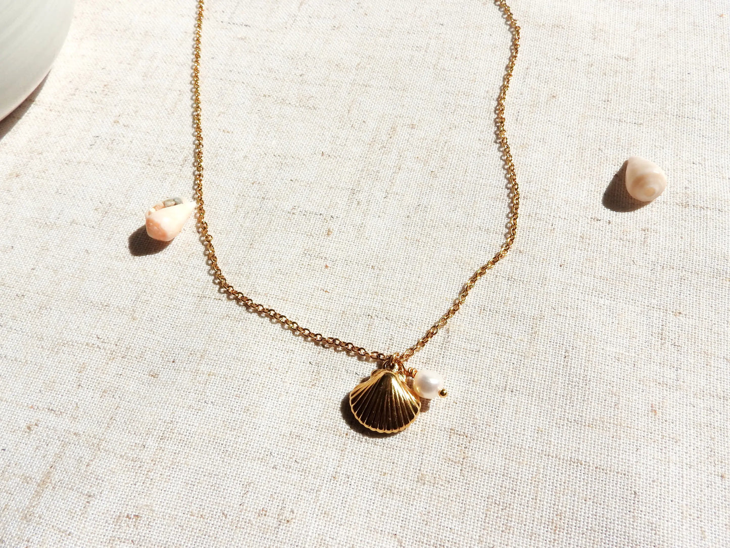 Collier chaîne fines mailles en acier inoxydable doré. Bijou orné d'un double pendentif coquillage en acier doré et une perle d'eau douce blanche. Bijou posé sur une nappe beige décorée de coquillages de mer.
