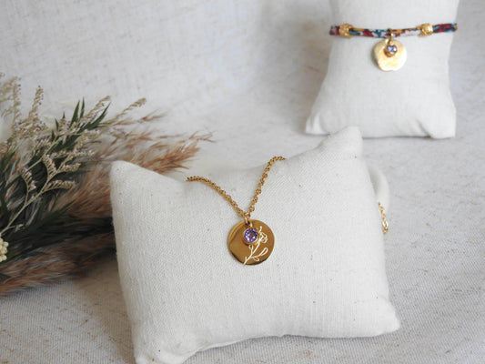 Collier chaîne fine avec pendentif fleur iris et sa pierre de naissance améthyste du mois de février. Bijou posé sur un mini-coussin beige en coton