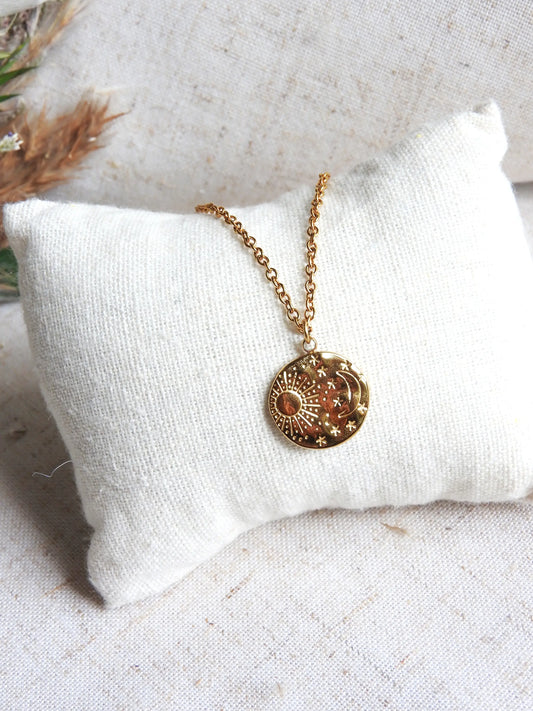 Collier chaîne pendentif médaille rond au motif astral avec le soleil, la lune et les étoiles. Bijou présenté sur un mini-coussin beige en coton.