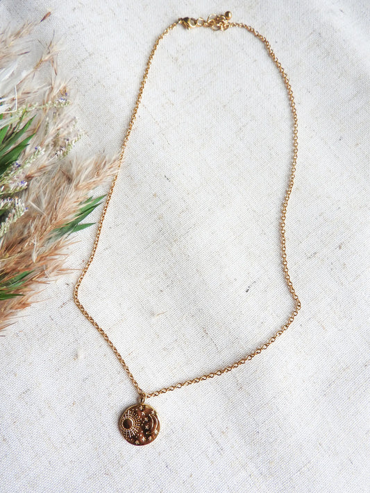 Collier chaîne pendentif médaille rond au motif astral avec le soleil, la lune et les étoiles. Bijou en acier inoxydable doré posé sur une nappe beige en coton décoré d'une brindille.