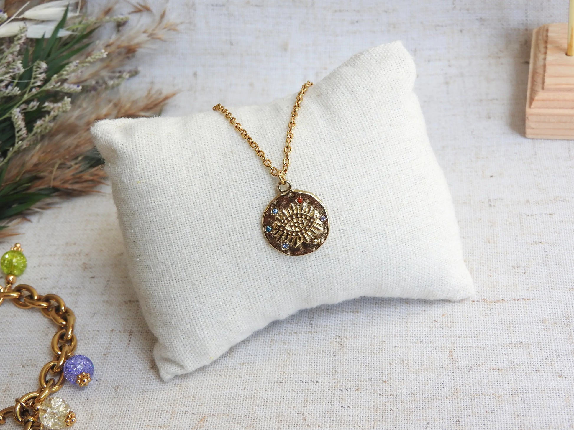 collier chaîne fine en acier inoxydable doré à pendentif médaille décoré d'un oeil protecteur et de strass multicouleurs. Bijou posé sur un mini-coussin beige, déposé sur une nappe décorée d'une brindille.