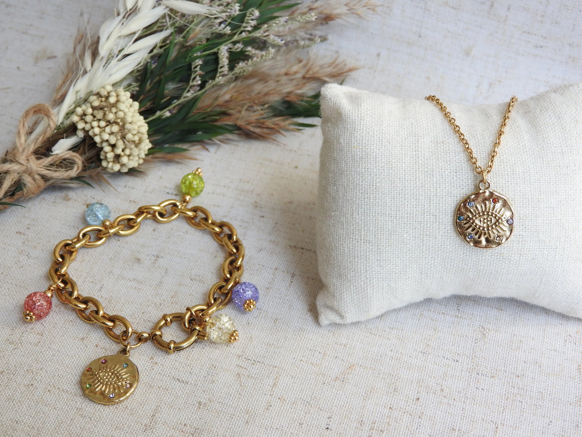 parure bijoux avec collier chaîne à pendentif rond orné de l'oeil protecteur et strass multicouleurs, bijou posé sur un mini-coussin beige et au côté d'un bralcelet à larges maillons en acier doré à breloques avec des perles de verre colorées et le même pendentif oeil protecteur.