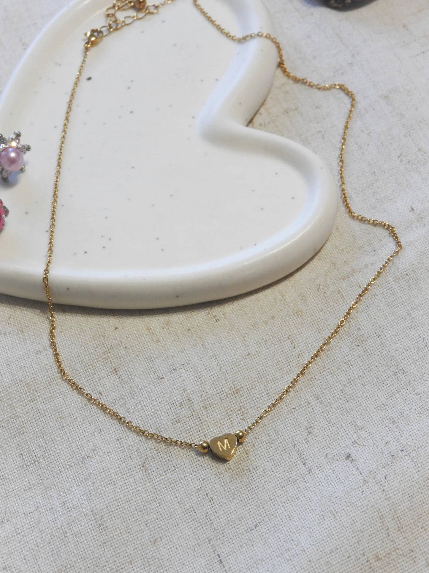 Collier chaîne fine en acier inoxydable doré posé à plat sur une nappe beige et une coupelle en céramique blanche. Bijou orné d'une perle plate en forme de coeur, gravée de la lettre M.