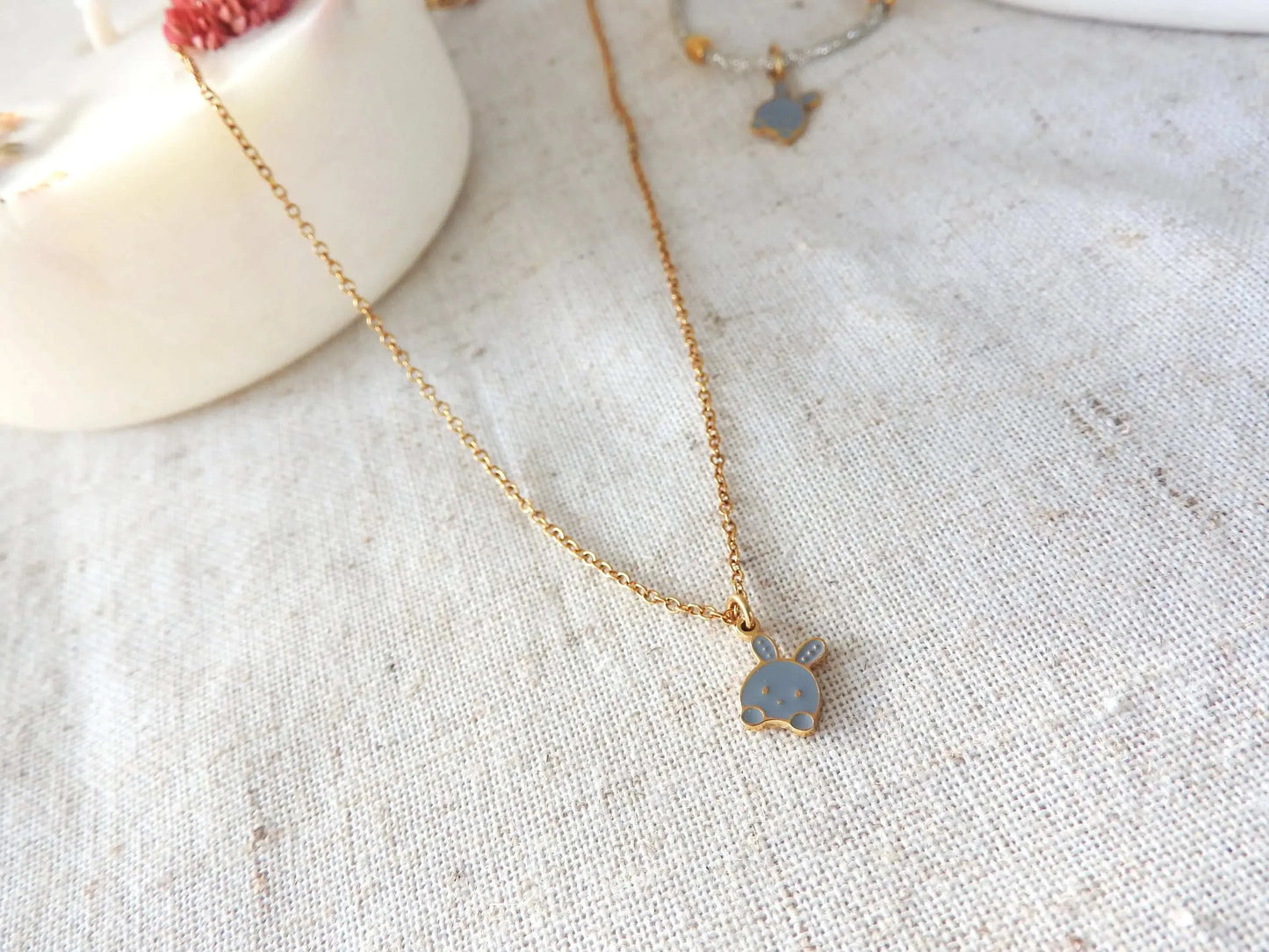 Collier enfant à chaîne fine en acier inoxydable doré et orné d'un pendentif lapin gris en résine époxy et acier inoxydable doré. Bijou posé à plat sur une nappe beige décorée d'une bogie fleurie.