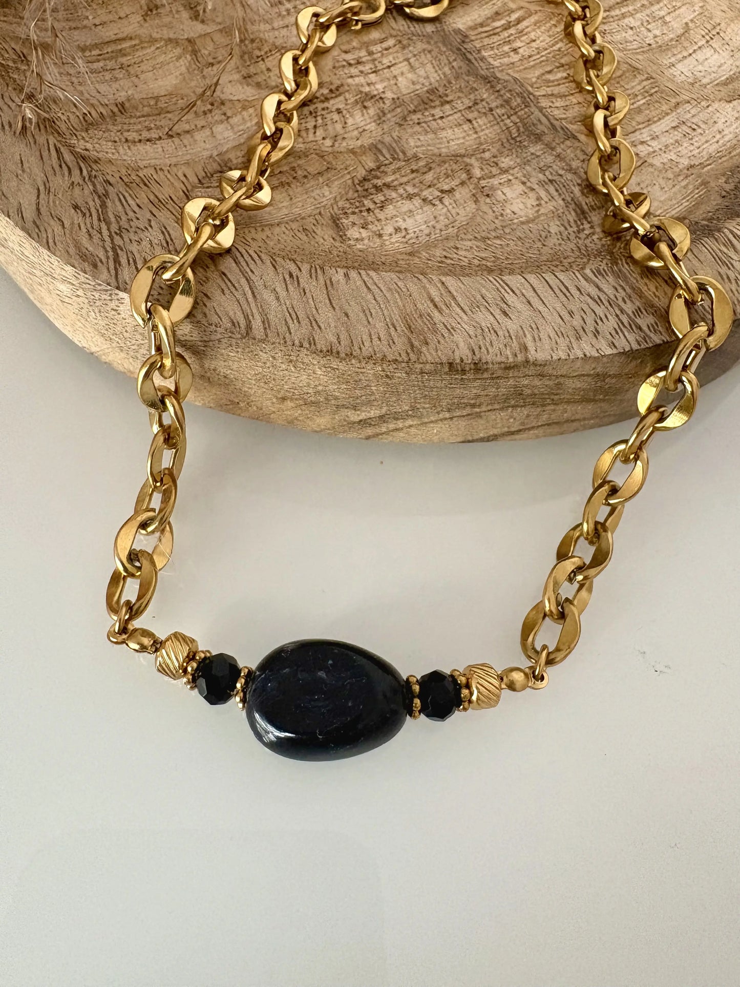 collier ras-du-cou à larges maillons en acier inoxydable doré, orné d'une grande perle irrégulière noire marbré. Bijou posé à plat sur une table en verre trempé et un plateau en bambou.