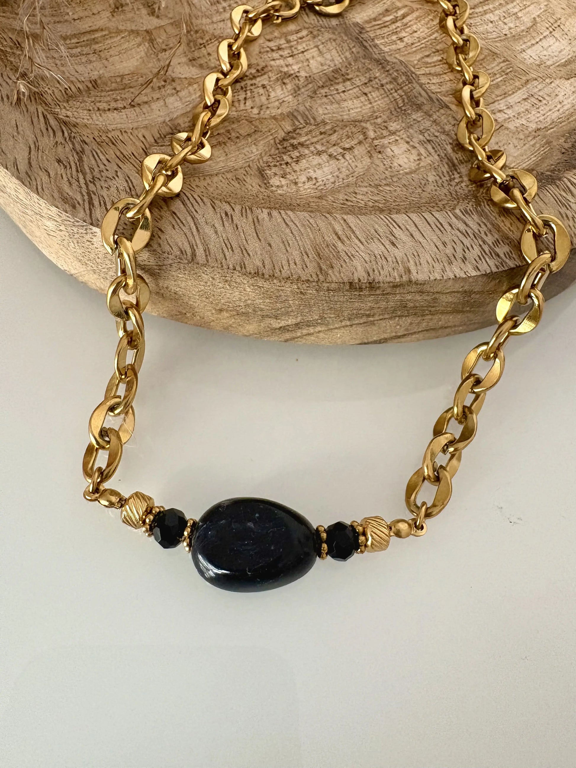 collier ras-du-cou à larges maillons en acier inoxydable doré, orné d'une grande perle irrégulière noire marbré. Bijou posé à plat sur une table en verre trempé et un plateau en bambou.