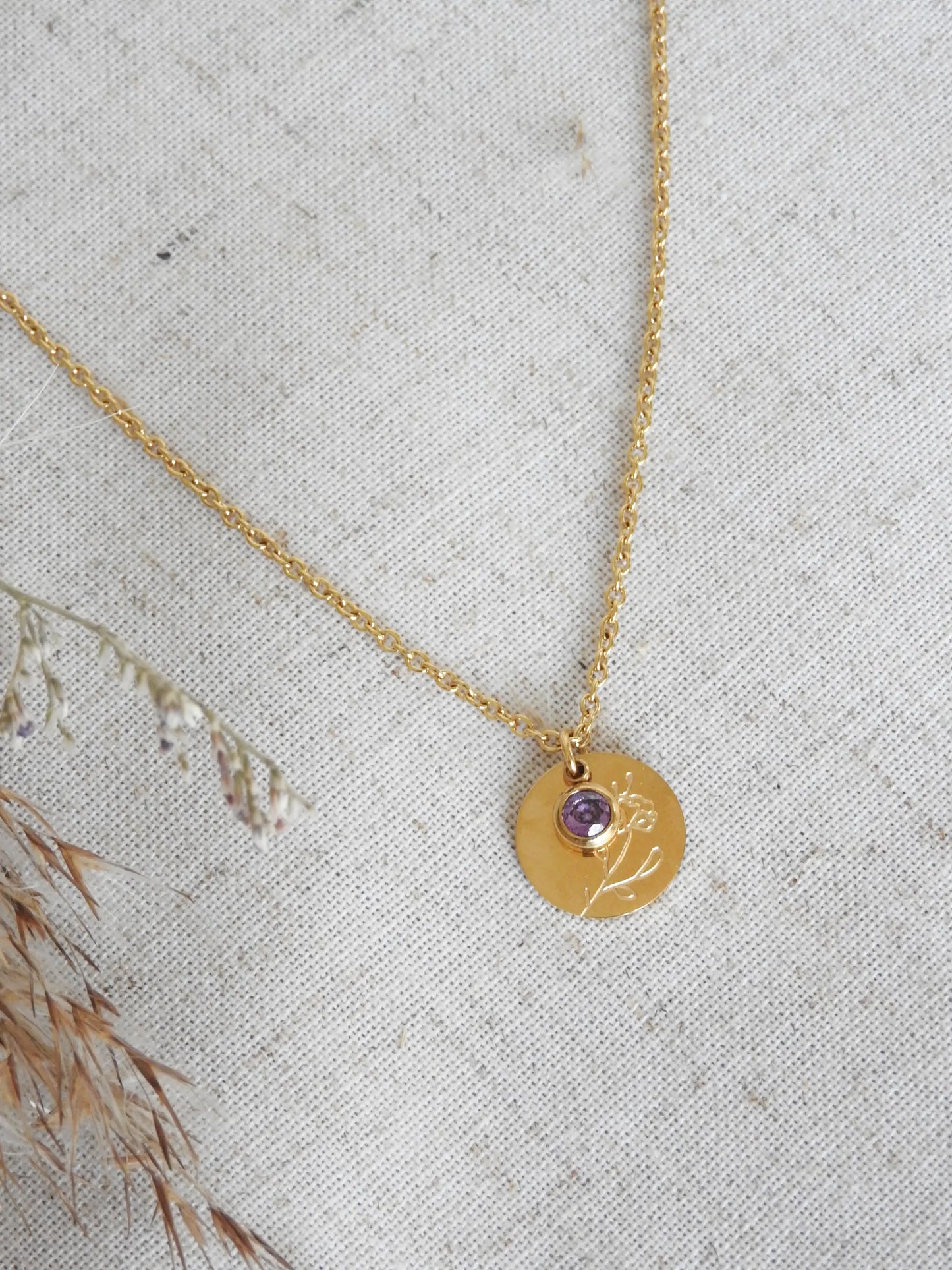 Collier chaîne fine avec pendentif fleur iris et sa pierre de naissance améthyste du mois de février. Bijou posé à plat sur une nappe beige en coton