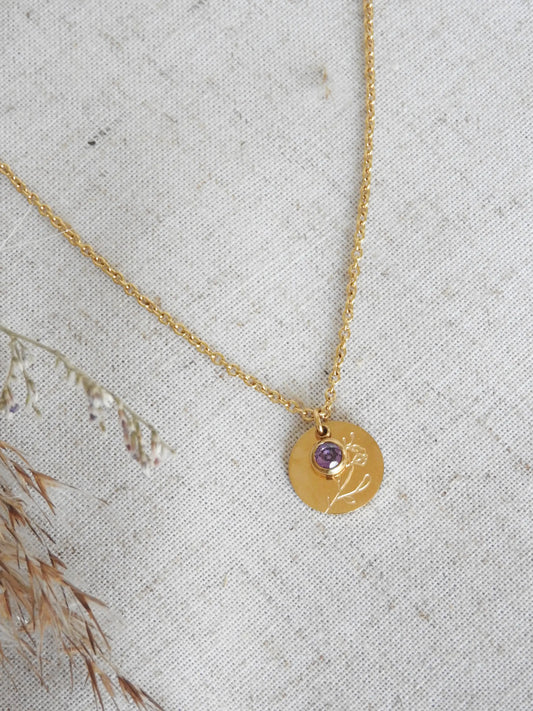 Collier chaîne fine avec pendentif fleur iris et sa pierre de naissance améthyste du mois de février. Bijou posé à plat sur une nappe beige en coton