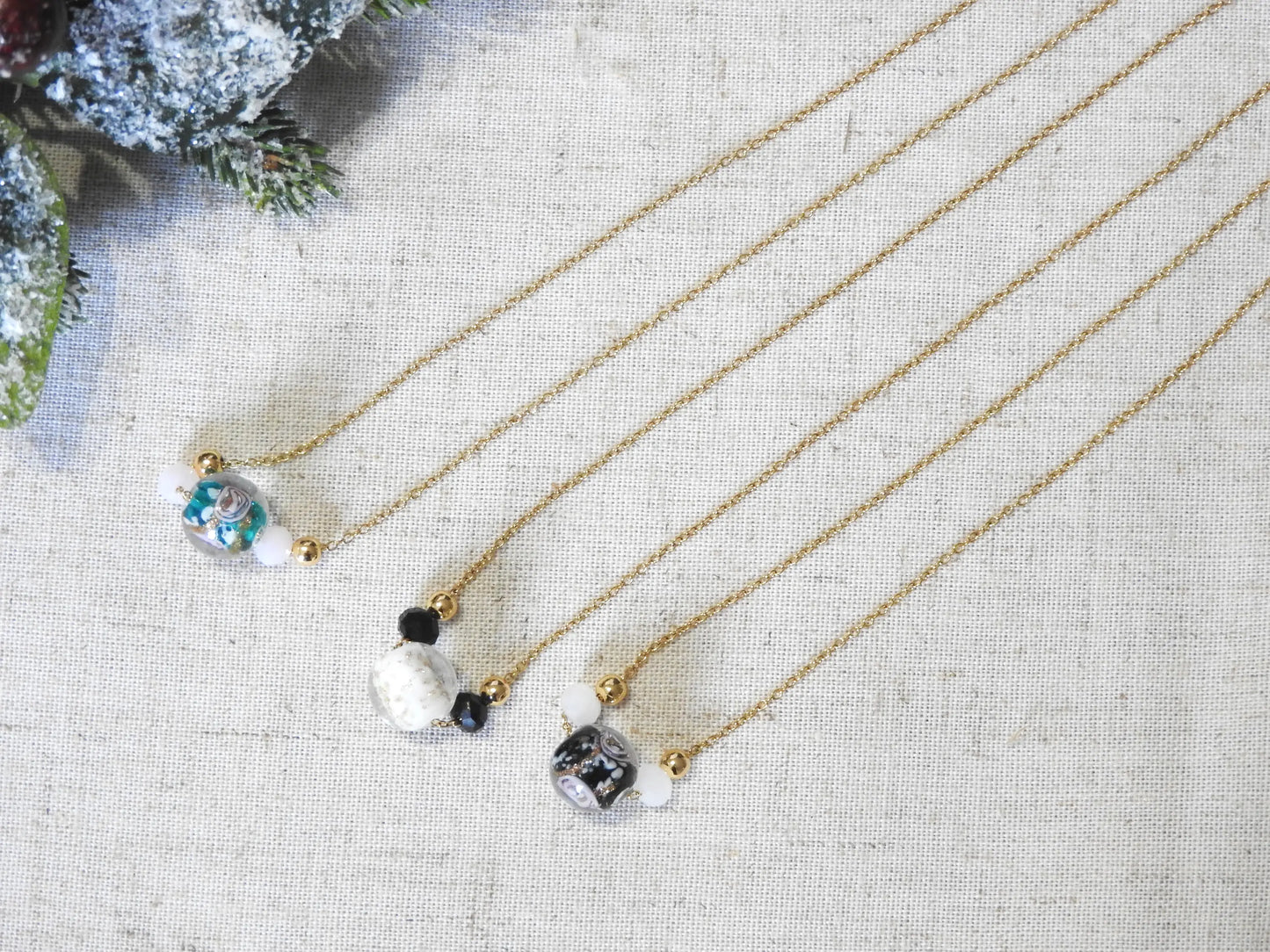 trois colliers posés à plat sur une nappe beige décorée d'une branche de sapin enneigée. Colliers chaîne en acier inoxydable doré, ornée de perles de verre et boule de verre à la décoration florale.