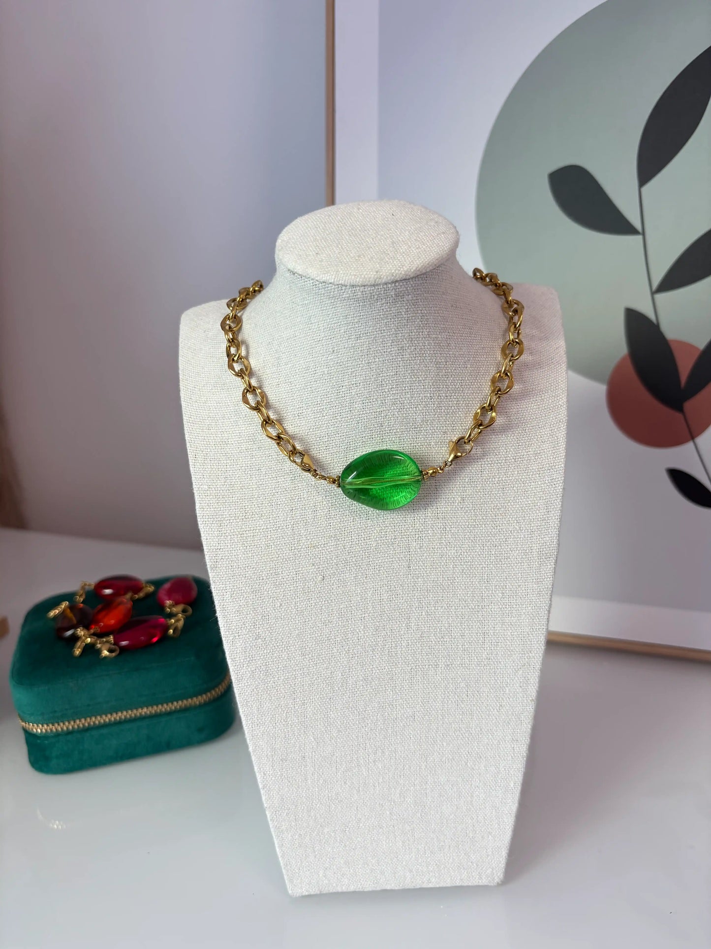 Collier à larges maillons en acier inoxydable orné d'une perle ovale irrégulière colorée verte translucide. Bijou présenté sur un buste en coton beige.
