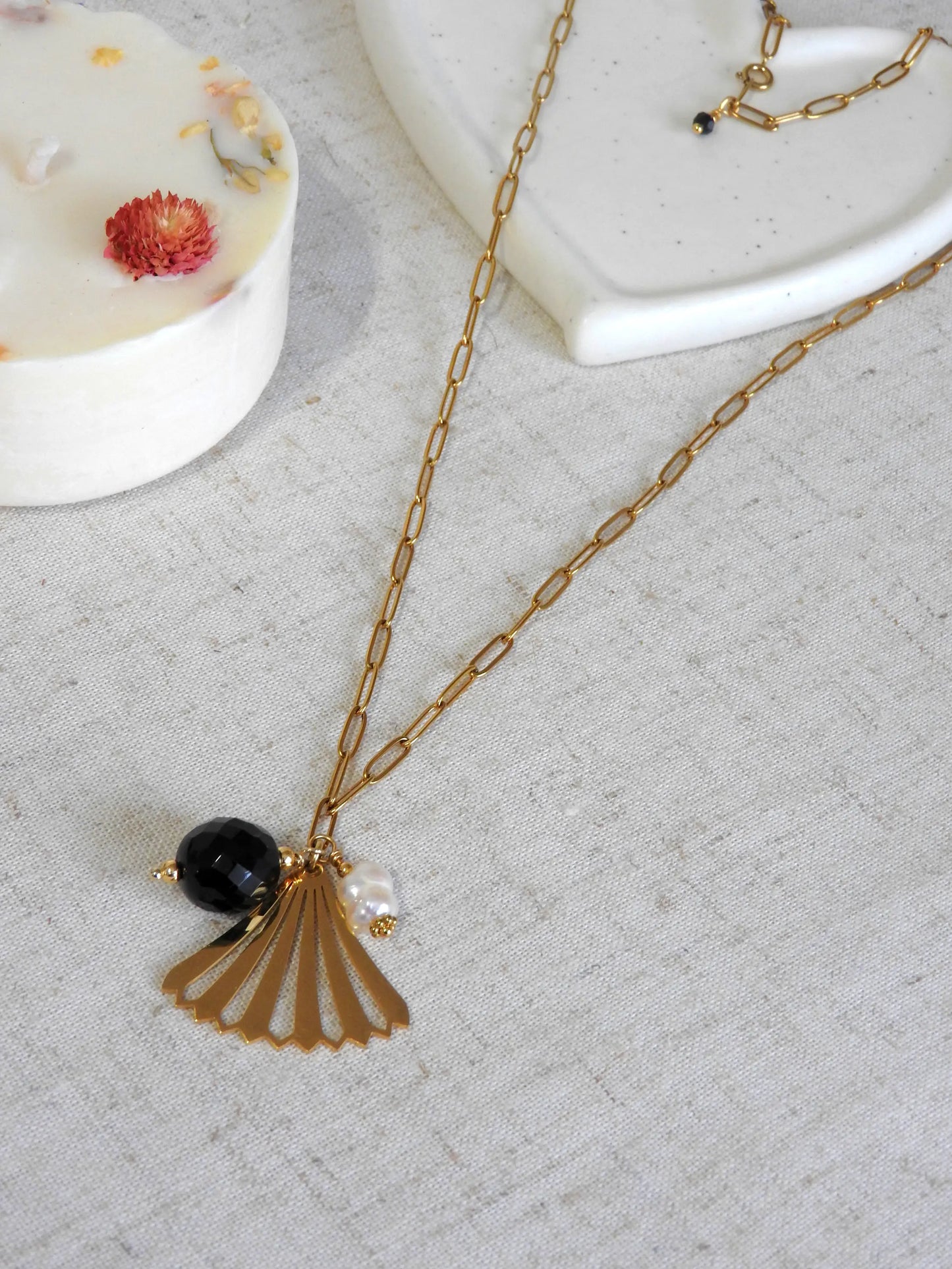 Collier sautoir en acier inoxydable doré, orné d'un pendentif en forme d'éventail doré, une perle onyx noire et une perle d'eau douce blanche.