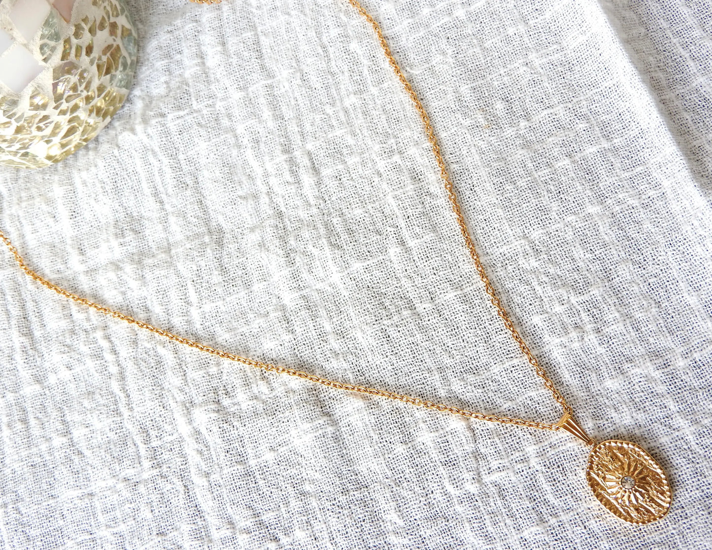 Collier sautoir chaîne en acier inoxydable doré avec une médaille ovale ornée d'un soleil en relief avec une pierre en oxyde de zirconium.  Bijou posé sur une nappe en gaze de coton blanche.