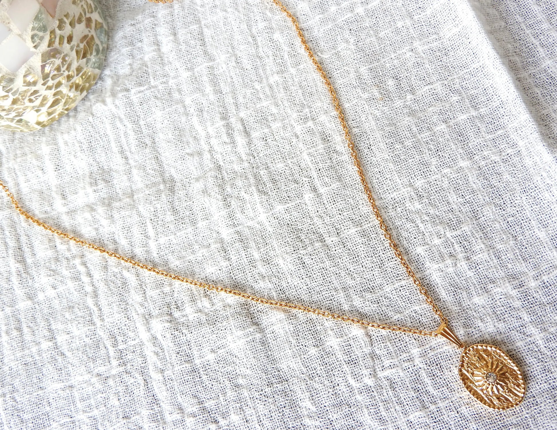 Collier sautoir chaîne en acier inoxydable doré avec une médaille ovale ornée d'un soleil en relief avec une pierre en oxyde de zirconium.  Bijou posé sur une nappe en gaze de coton blanche.