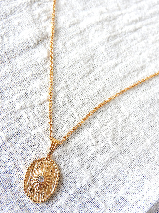 Collier sautoir chaîne en acier inoxydable doré avec un pendentif médaille de forme ovale orné d'un soleil en relief avec une pierre en oxyde de zirconium.  Bijou posé sur une nappe en gaze de coton blanche.