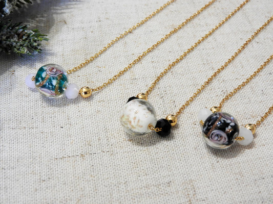 trois colliers posés à plat sur une nappe beige décorée d'une branche de sapin enneigée. Zoom sur les colliers ornées de boule de verre au motif floral de couleur verte, blanche et noire. Bijoux 
en acier inoxydable doré.