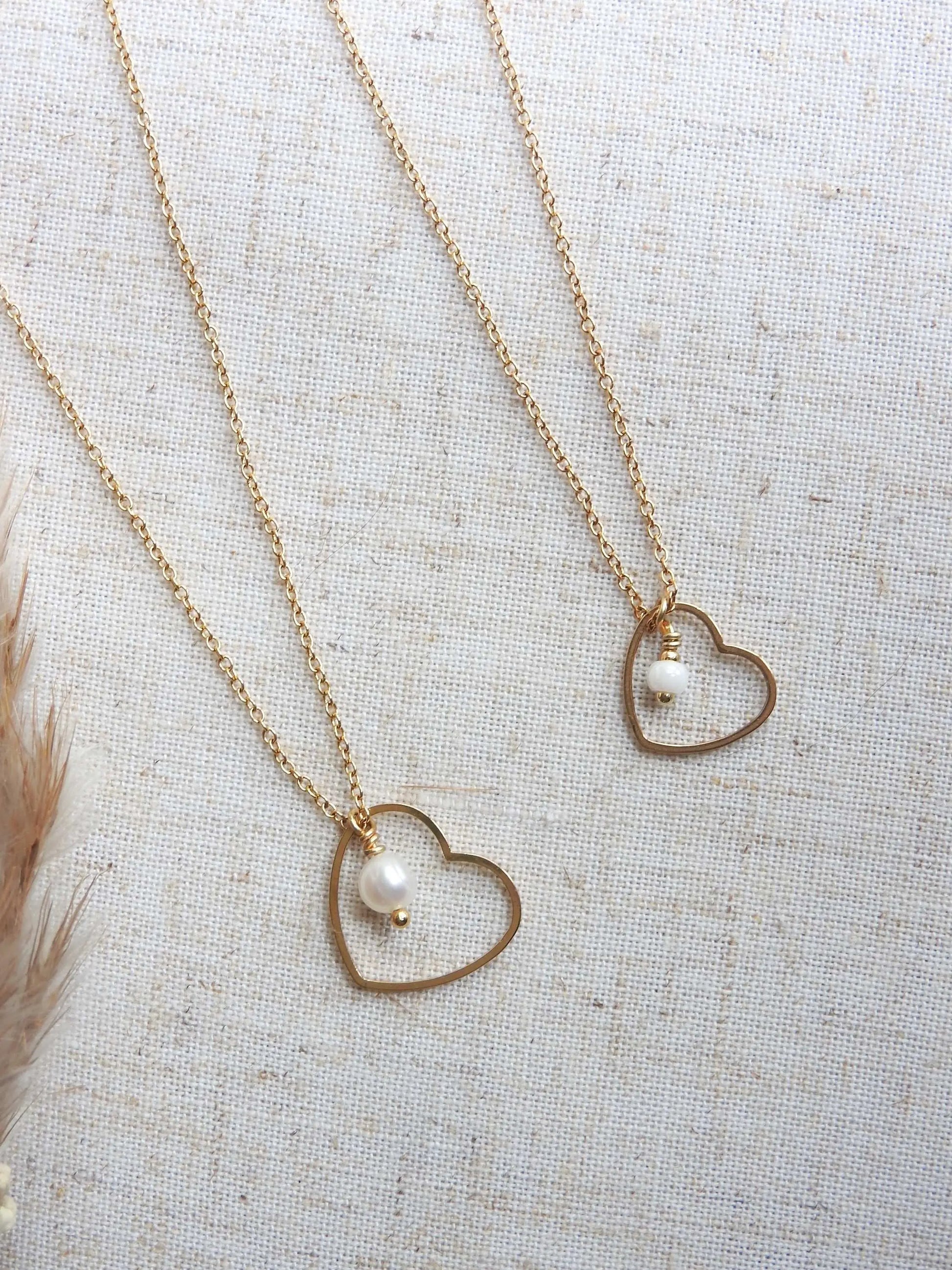 deux colliers côte à côte posés à plat sur une nappe beige, chaîne fine en acier inoxydable doré avec un pendentif coeur évidé et orné l'un avec une perle d'eau douce et une perle de verre pour le 2ème collier.