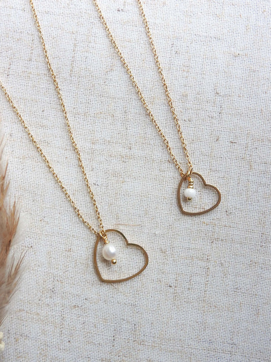 deux colliers côte à côte posés à plat sur une nappe beige, chaîne fine en acier inoxydable doré avec un pendentif coeur évidé et orné l'un avec une perle d'eau douce et une perle de verre pour le 2ème collier.