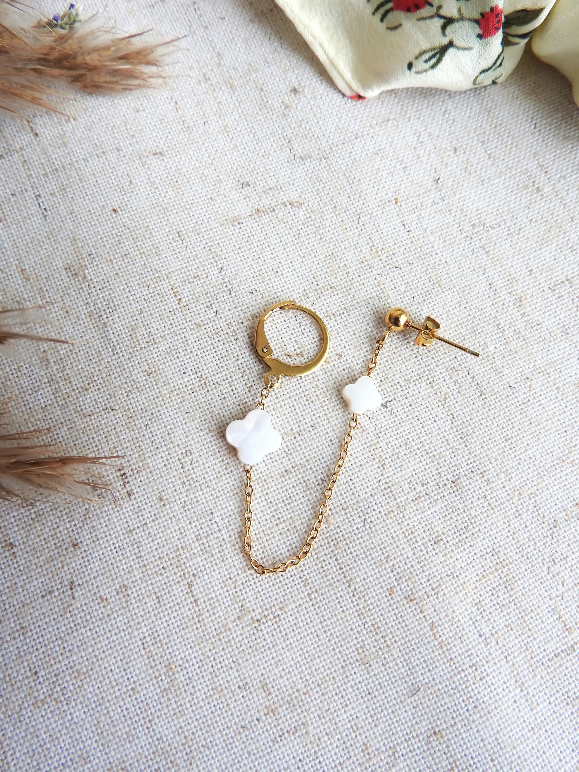 Double boucle d'oreilles à clou boule et une mini-créole reliée par une chainette ornée de deux perles trèfle en nacre blanche et en acier inoxydable doré. Bijou posé sur une nappe beige décorée d'une brindille.