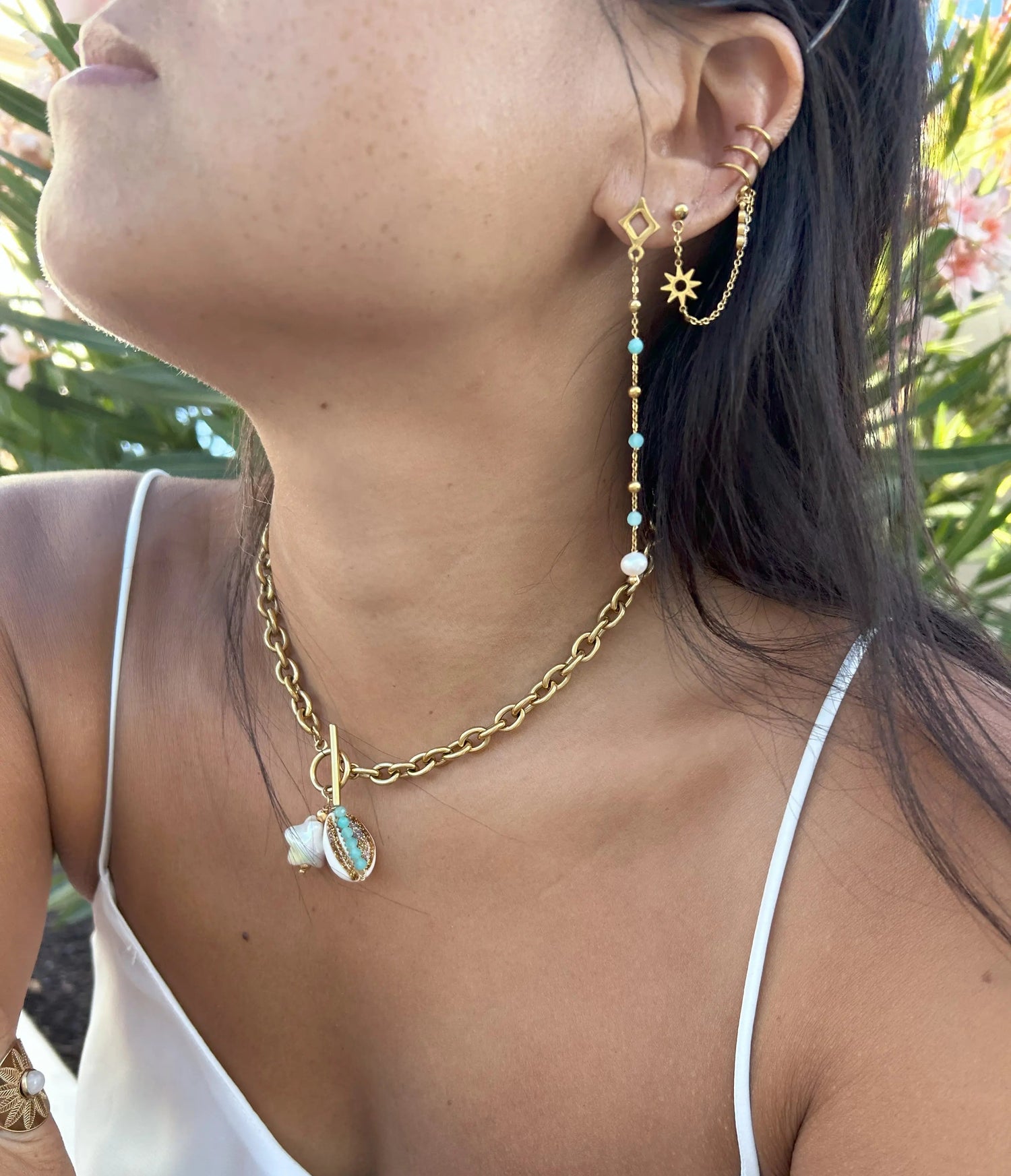 femme portant des bijoux en acier inoxydable doré, collier grosses mailles avec breloques coquillage cauri et étoile en acrylique blanc, boucles d'oreilles chaines pendantes perlées en acier doré et en verre turquoise et perle d'eau douce. Triple bague d'oreille avec barre scintillante reliée à une chaînette avec un intercalaire soleil.