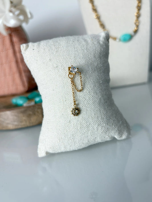 mono-boucle d'oreille chainette en acier inoxydable doré avec clou décoré d'une pierre en oxydes de zirconium et un mini-soleil strass suspendu à la chaînette. Bijou présenté sur un mini-coussin en coton beige.