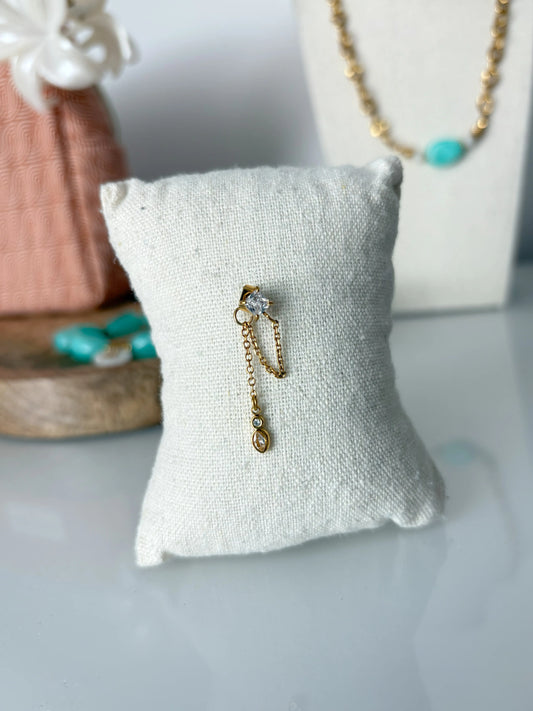 Mono-boucle d'oreille chaînette en acier inoxydable doré avec clou et goutte en oxyde de zirconium. Bijou présenté sur un mini-coussin en coton beige posé sur une table décoré en fond d'un buste présentoir et un plateau avec une trousse de toilette.